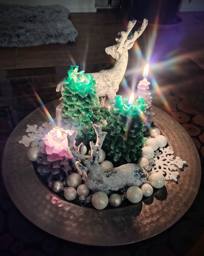 Ein Silberteller auf dem zwei weiße Dekohirsche, weiße und silberne Christbaumkugeln, weiße Kunststoffschneeflocken und vier brennende Kerzen in Nadelbaumoptik zu sehen sind.