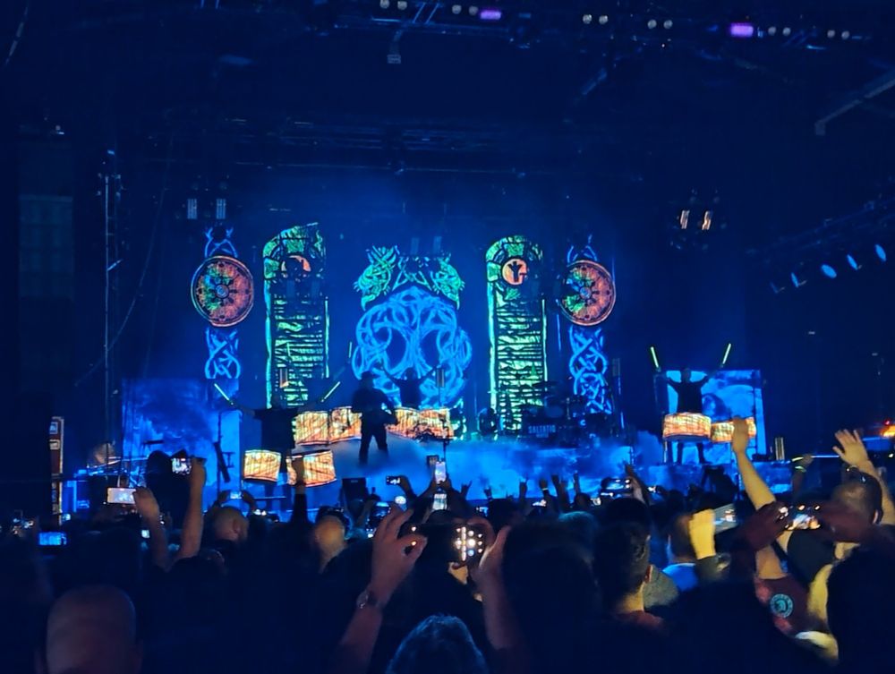 Saltatio Mortis Konzert Bühne:
Schwarzlicht-Beleuchtung, grün-blauer Hintergrund, orange-gelbe Trommeln im Vordergrund, die Band trägt schwarz und passende Neonfarbene Drumsticks