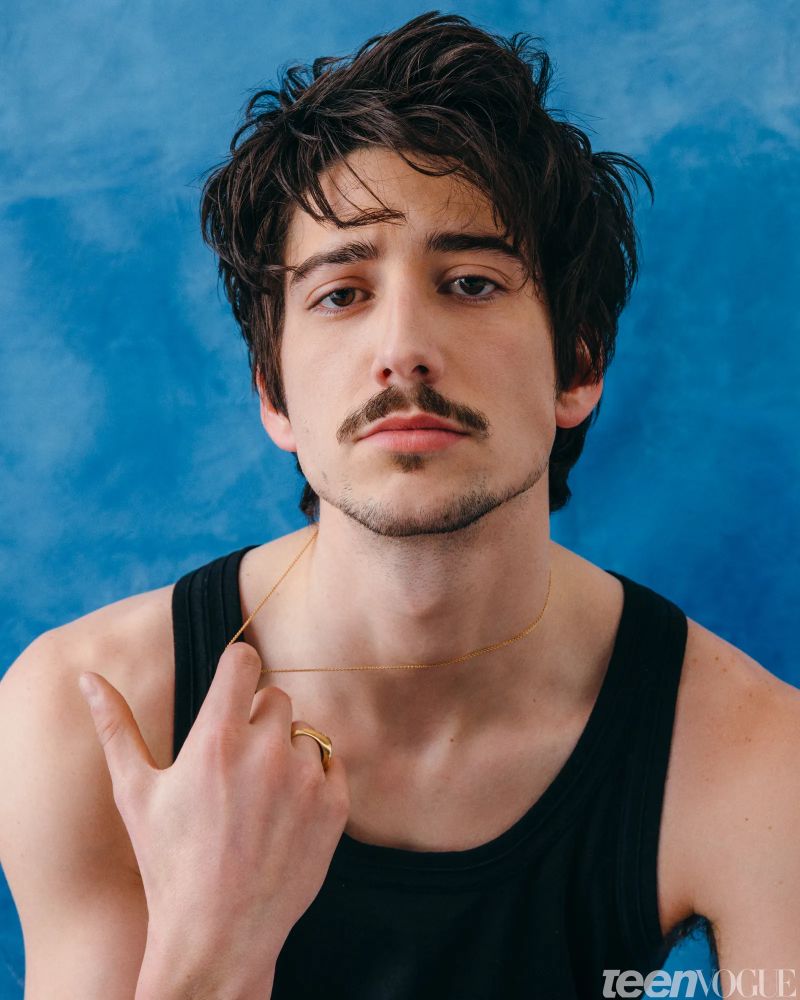 Milo Manheim in a black tank top pulling on a small chain that’s around his neck