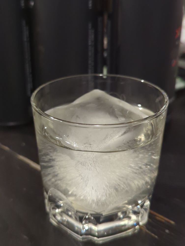 A whiskey glass containing one large ice cube and onionade. The onionade is off white and slightly cloudy. 
