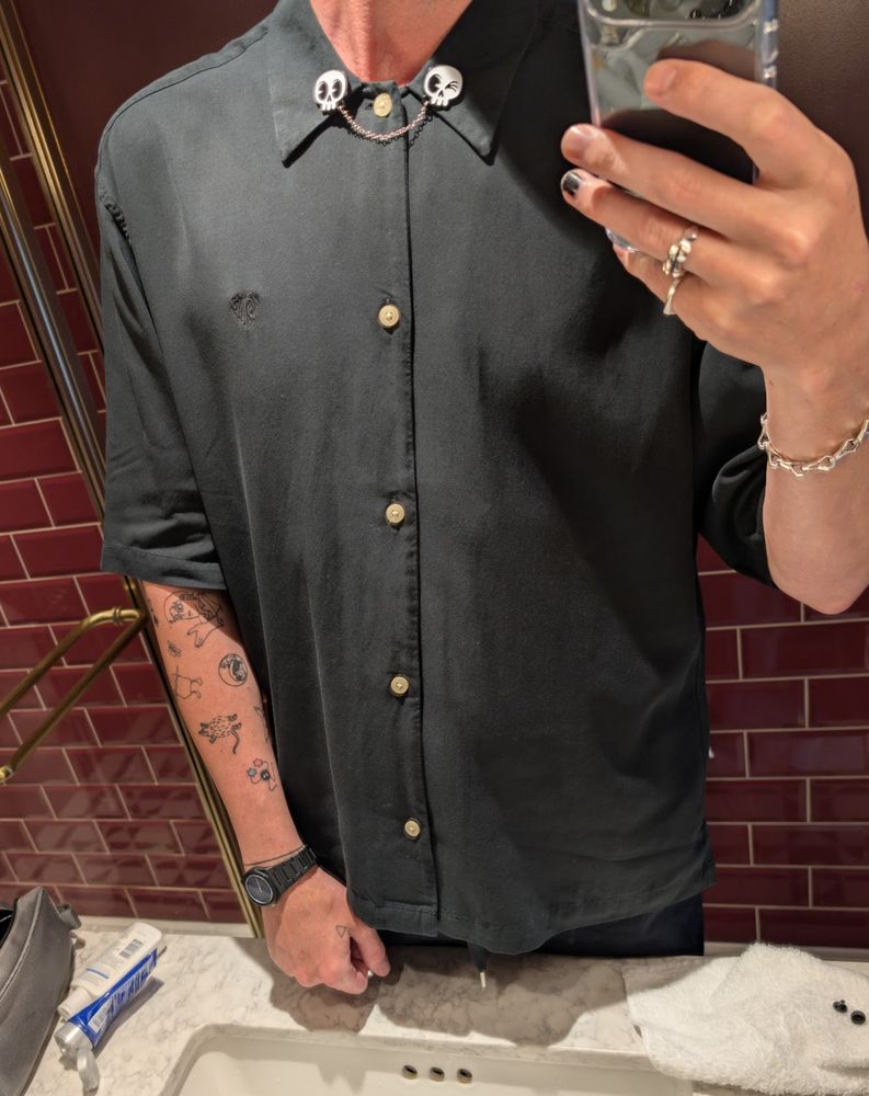 mirror selfie of some dood wearing an oversized black short sleeve button up with a pair of skull enamel pins linked by a chain attached to the collar