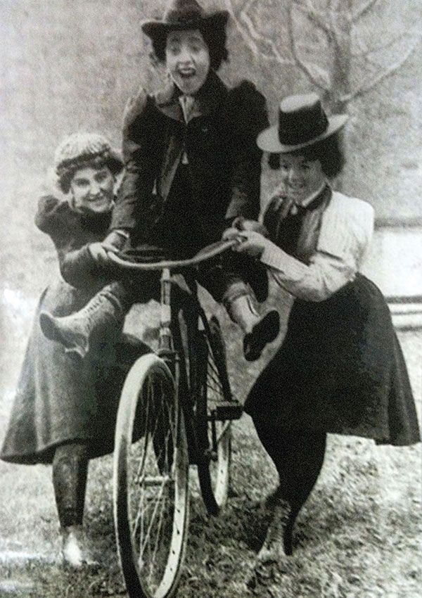 Image shows a young Victorian woman wearing a form of bloomers, being gleefully pushed on her bike by female friends, dressed in more conservative clothes