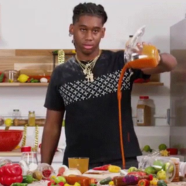 a man is pouring orange juice into a glass while standing in a kitchen surrounded by vegetables .