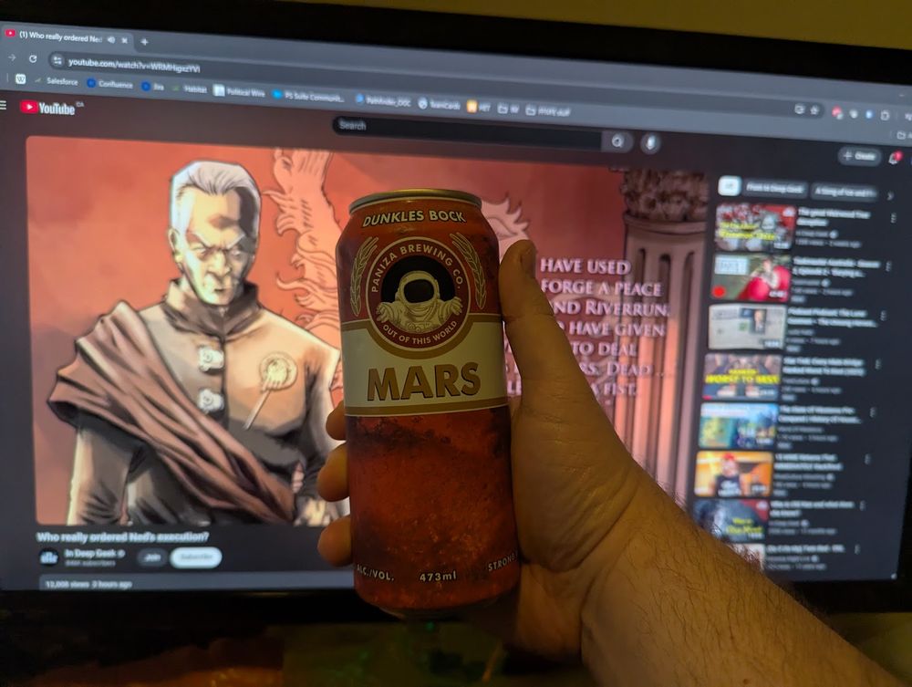 My right hand, holding a tallboy of Paniza Brewing Co Mars Dunkels Bock. The beer has an image of the Martian surface on the can and the logo is an astronaut in a red window surrounded by laurels. A monitor showing a youtube video from In Deep Geek is in the background.