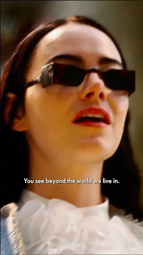 A screenshot from the movie Poor Things. Emma Stone, mouth slightly open, wearing futuristic rectangle sunglasses. Words displayed, read ‘You see beyond the world we live in’