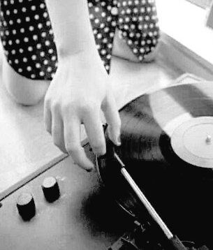 A figure wearing a polka dot dress appears only from waist down. Kneeling in front of an old time record player gently holding the needle above the album . 