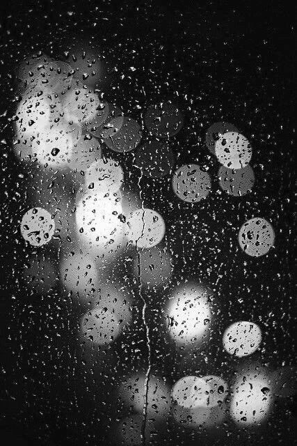 A pane of glass speckled with delicate raindrop highlighted by street and traffic light In black and white 