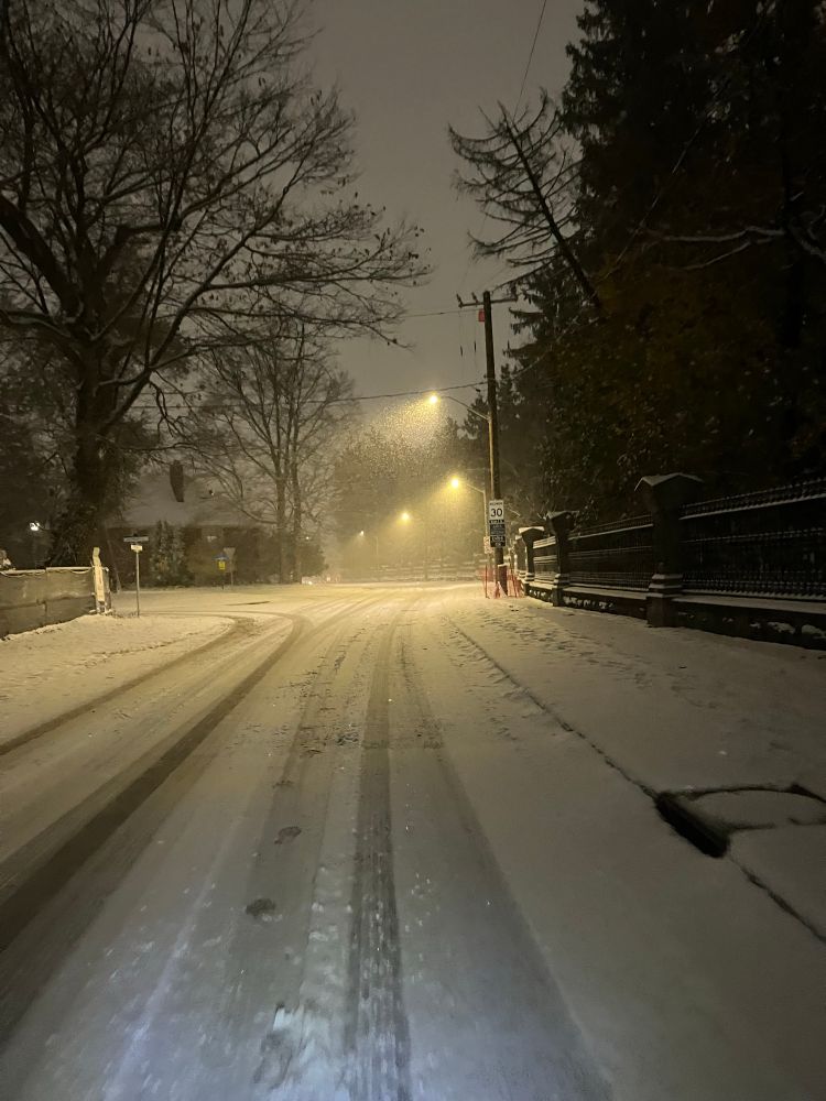 Road at night covered in fresh snow. One can see car tire tracks and foot prints. Street light in the background and the Governor General’s property to the right. 