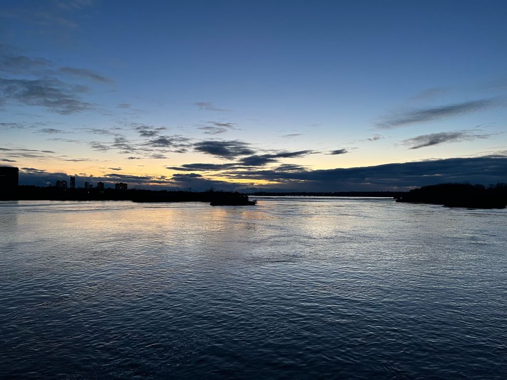 View of the Ottawa River as seen from the Chief-William-Commanda bridge. One can see a skyline on the left, an island in the background as well as the shoreline on the right. Some clouds on the horizon and a skyline that goes from yellow to light blue at the top. 