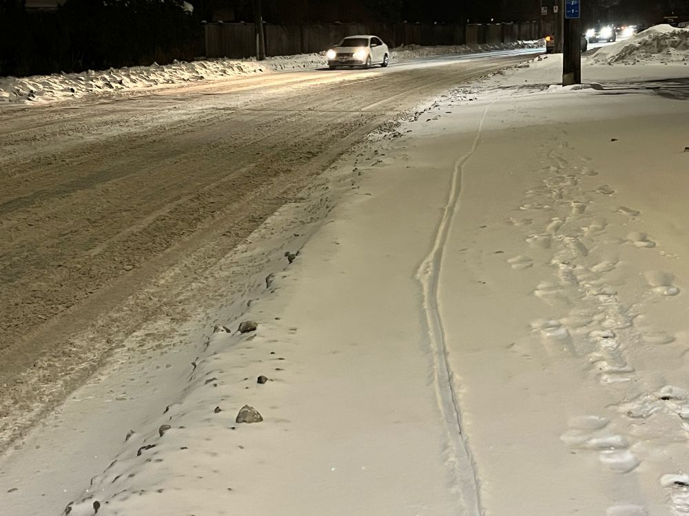 A road in a snow storm. The road is covered in slosh, as is the painted bike lane. The sidewalk on the right is covered with a layer of fresh paint. There are two bike tire tracks in the snow. 