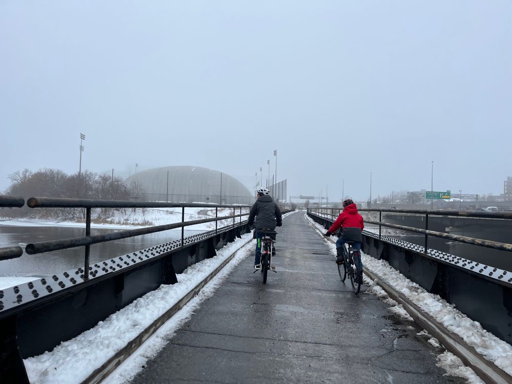 Parent and a child biking on a former railway bridge on a foggy day. There’s some snow on the sides and on the river’s edge. 