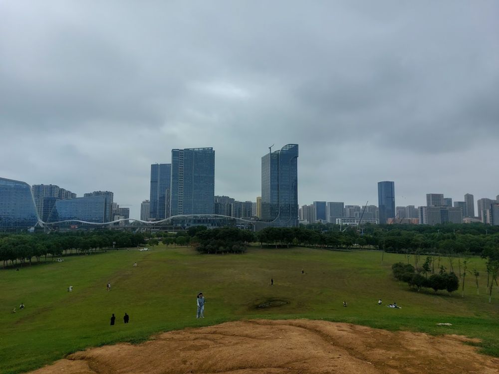 A city scene taken from a park, so there's a wide green expanse with trees in the foreground and many skyscrapers in the background. There are a few people dotted in the distance, walking or lying in the grass together. Then there's a child who looks like she's also far away but just like 3 × the size of the other people