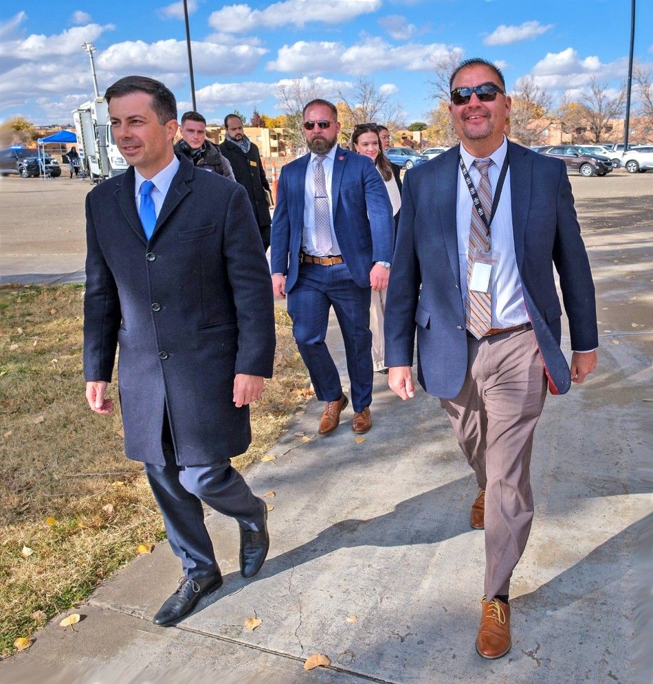 From the Los Alamos Daily Post, an outdoor photo with a few stray leaves on the ground and light coats, with Pete Buttigieg walking in appropriate clothing, shoes, and socks next to the New Mexico Transportation Secretary, Richey Serna, followed by others. "U.S. Secretary of Transportation Pete Buttigieg, left, walking with New Mexico Department of Transportation  Secretary Ricky Serna, right, during their visit Wednesday to Southwestern Indian Polytechnic Institute (SIPI) in Bernalillo County. Secretary Buttigieg made a special trip to New Mexico to meet with tribal leaders, students and others and conduct discussions on roadway safety and improvements to local infrastructure. Courtesy photo"