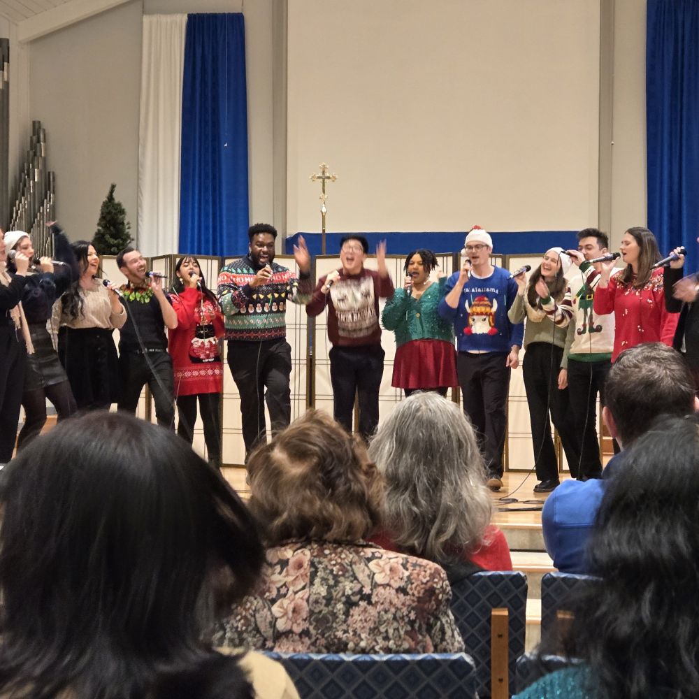 A choir group performing in a church. 11 or 12 singers are visible, singing into handheld microphones. They are wearing various ugly Christmas sweaters. Blue and white curtain tapestries hang in the background. The backs of the heads of audience members are visible in the foreground.