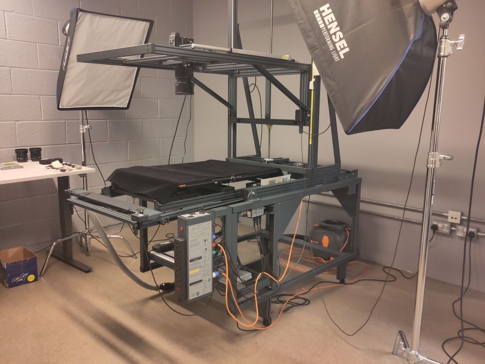 Large metal frame with a black flat area to hold a book, a camera suspended above it, and two huge lights aimed at it.