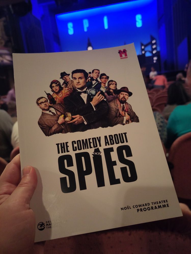 A hand holding a theatre programme for The Comedy About Spies. Behind the programme you can just see an audience and a blue stage.