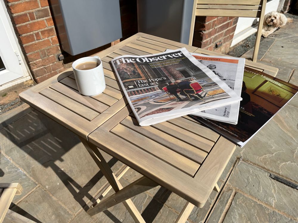 The Observer newspaper spread out on the little folding table outside my back door, with a mug of coffee and small hairy dog photobombing in the top corner of the picture 