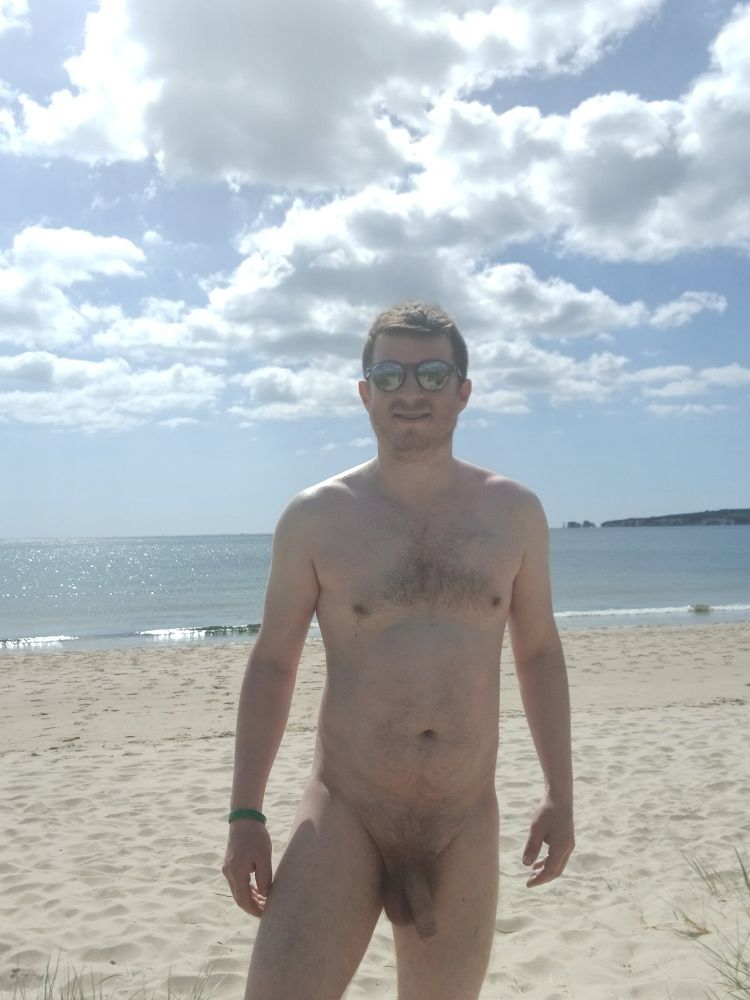 Unclothed white male facing the camera with glasses on, with all on show on the sand at a beach on the coast with sea, mostly blue sky and some clouds in the background 