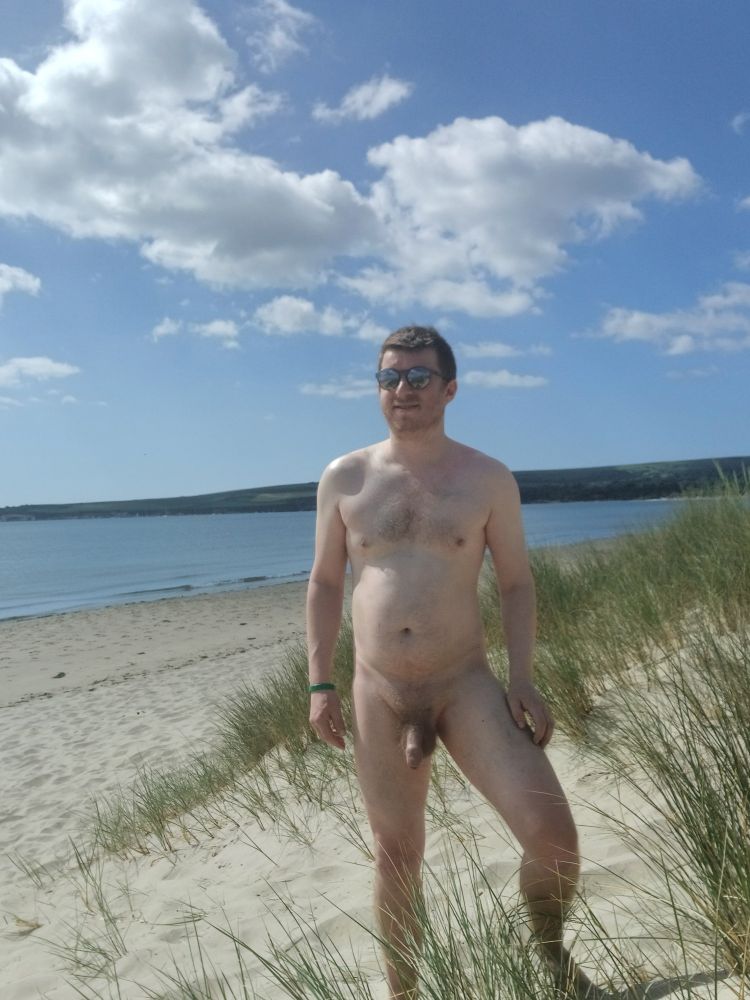 Naked white man on a beach enjoying the sun with reeds to the right and more land beyond the water. 