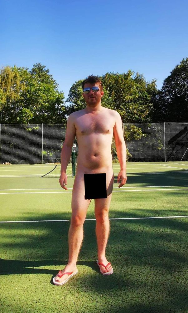 White male posing without clothes, only with flip flops and sunglasses on a tennis court in a naturist club with trees in the background and perfect blue sky 