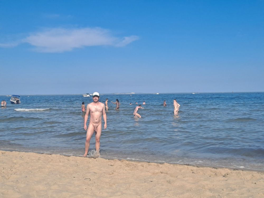 Full frontal unclothed white male at naturist beach with sand in foreground and sea and blue sky in the back with other naturists vaguely in the background. 