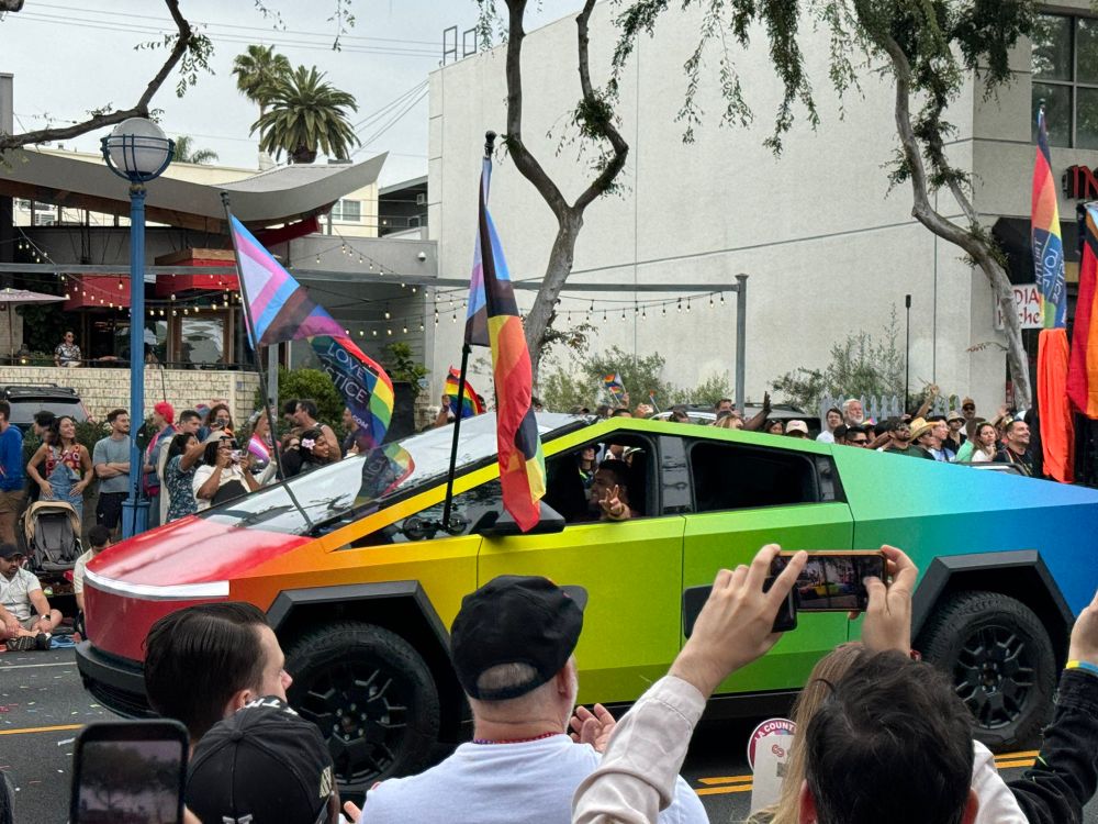 A Tesla Cybertruck wrapped in a rainbow gradient design, from red at the front to blue at the back, drives in a crowded Pride parade.
