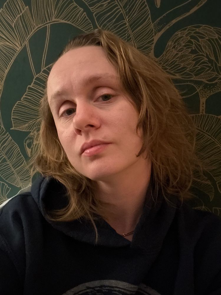 A tired white person in a hoodie gazes at the camera while leaning their head against a wall behind them with green wallpaper 