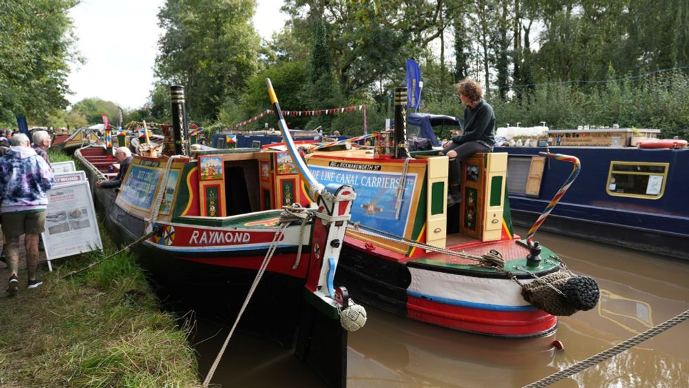 Two historic narrow boats moored at the side of a canal, with a trading boat on the other side of the canal nd people walking along the towpath.  
