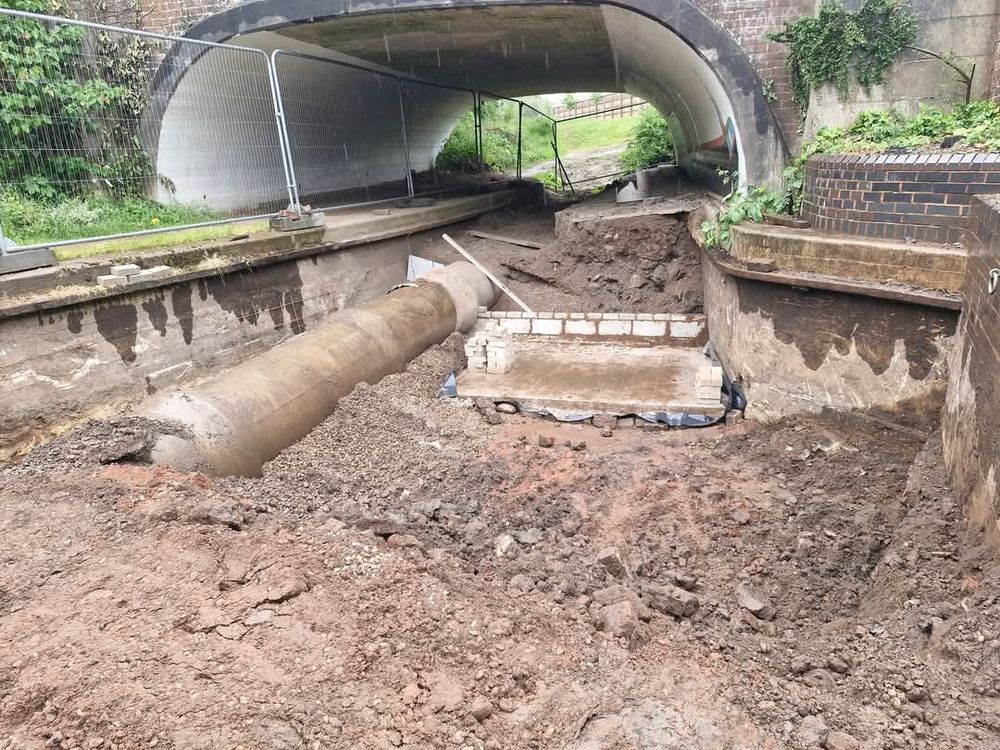 An empty canal channel that leads under a road bridge. The sides of the canal are original and there is a big pipe in the bed of the channel. There is a new wall of concrete blocks being built across the canal. 