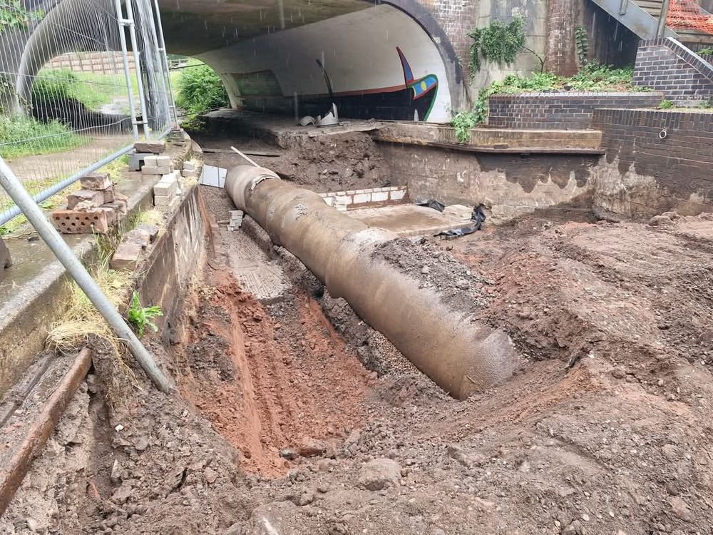 An empty canal channel that leads under a road bridge. The sides of the canal are original and there is a big pipe in the bed of the channel. There is a new wall of concrete blocks being built across the canal. 