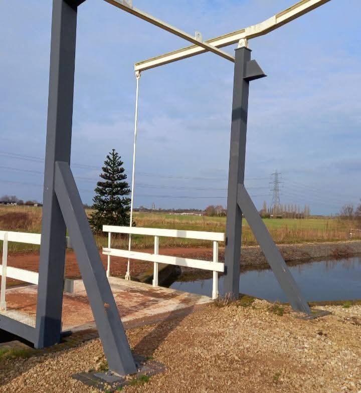 A close-up photo of a black and white liftbridge over a canal, with a fir tree in the background and a blue sky, with some wispy clouds.