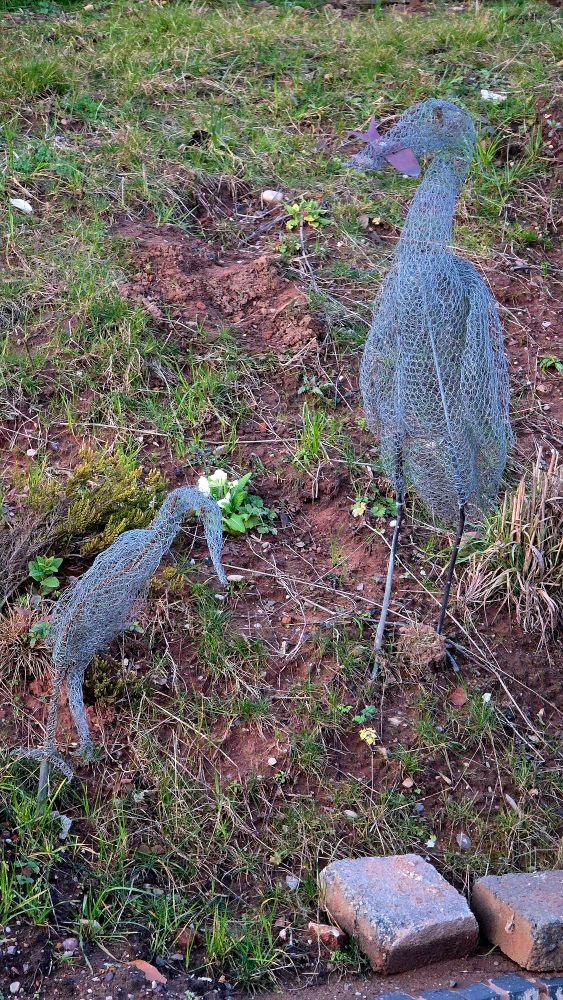 Two wire sculptures of herons on a canal bank. The larger one has a metal fish in its beak.