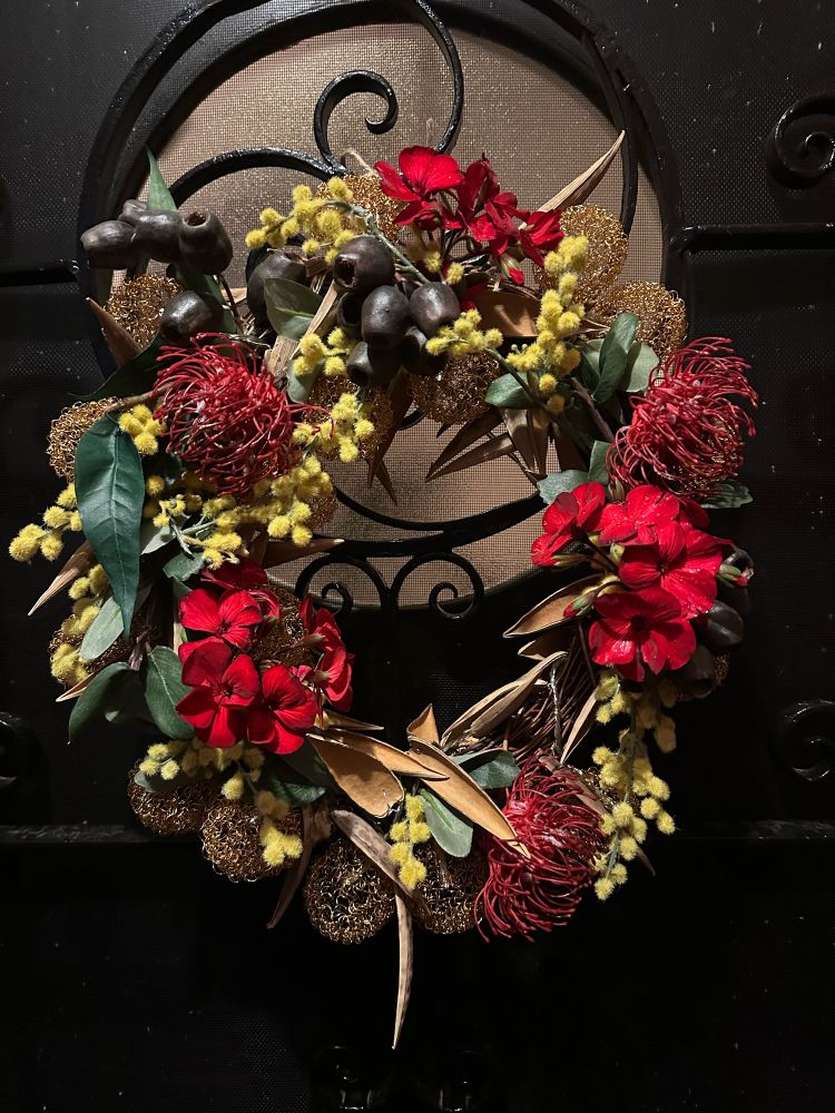 Christmas wreath with wattle, gum nuts, gum leaves made from wood, red geranium and lights hanging on front  door .