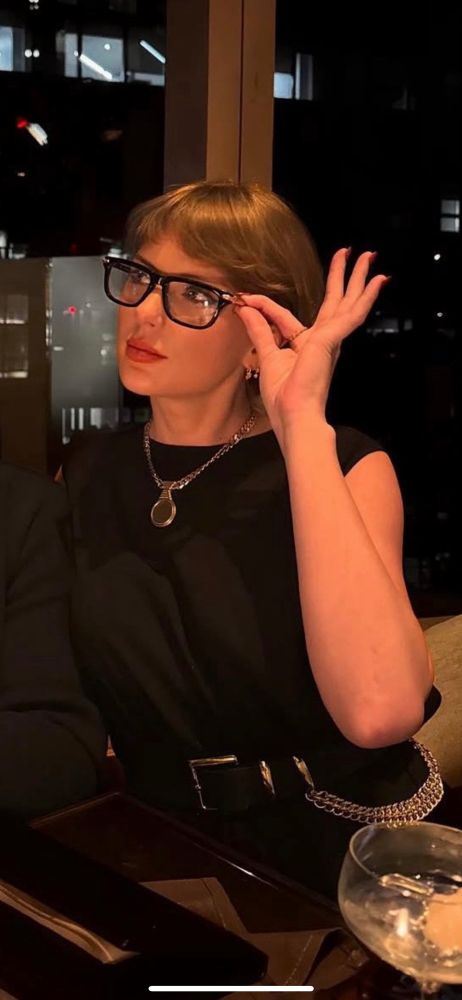 taylor swift wearing glasses with a think black frame, holding them with one hand. taken from travis’ instagram post