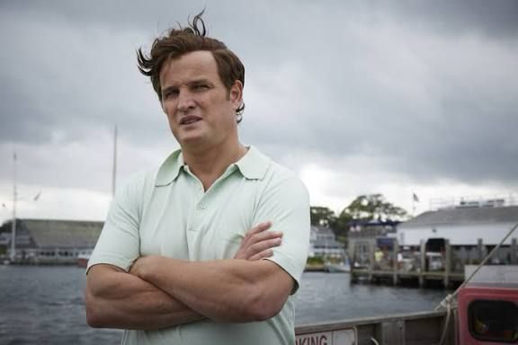 A man in a polo shirt stands with his arms folded with a harbour behind him. 