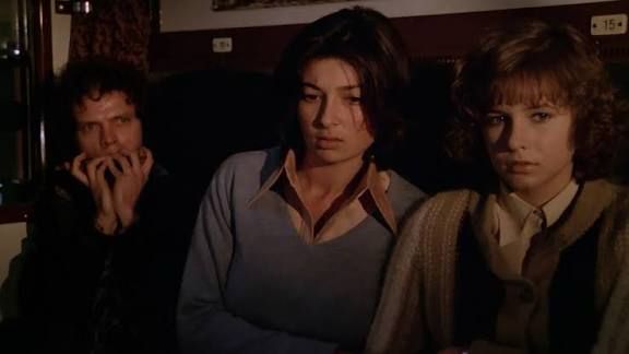 Two young women sit on a train next to each other. At the left side of them is a young man playing a harmonica. 