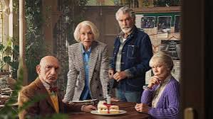 A group of pensioners two men and two women are round a table with a cake in it. The man on the left is seated, as is the woman on the right. Next to the seated man is a woman leaning forward onto the table. The fourth man is stood to her left.