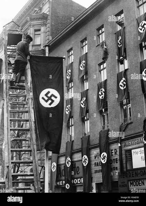 A horrible black and white photo of a street in Nazi Germany bedecked with Nazi flags from every apartment window and a Nazi flag being attached to a lamppost. 