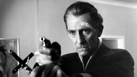 A man with a plaster over his left eyebrow holds a cross in one outstretched hand and a small gun in the other rested on top of the hand holding the cross.