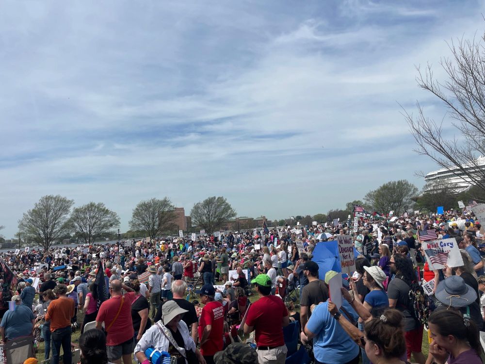 A massive crowd of people gathers outdoors, holding signs, under a cloudy sky, with trees and buildings in the background.