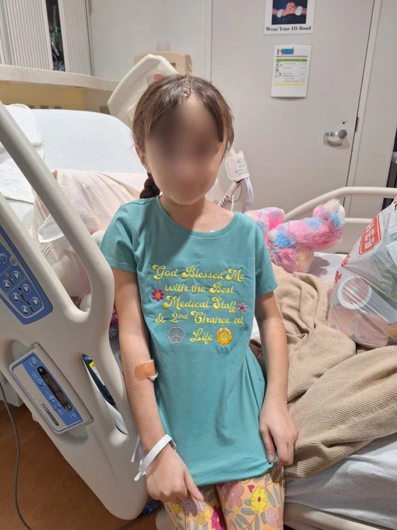 A child in a hospital bed, wearing a teal shirt with an uplifting message, surrounded by soft toys and cozy blankets.