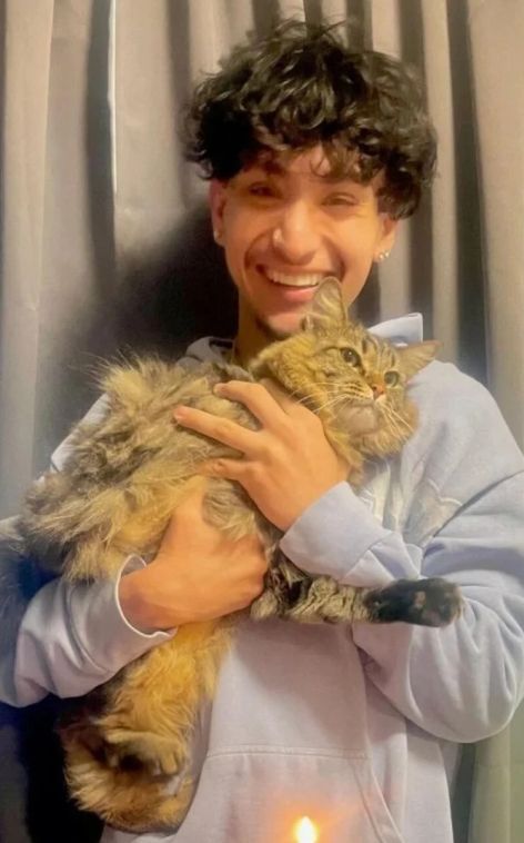 A smiling young man in a hoodie cradles a fluffy tabby cat against a backdrop of soft curtains and a candle.