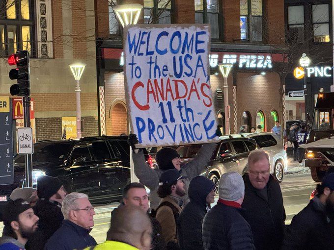 A protester holds a large sign reading "WELCOME to the U.S.A. CANADA'S 11th PROVINCE," amidst a crowd at night near storefronts.
