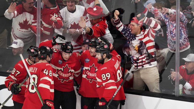 A group of Canadians hockey players in red jerseys celebrating, with enthusiastic fans in the background, some wearing patriotic US clothing while booing.