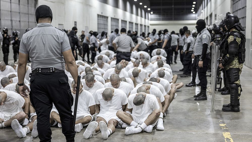 A picture of victims of ICE, all men on their knees with shaved heads bowed in front of sadistic, looking men with sticks and guns