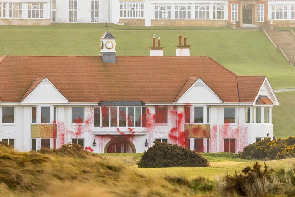 A vandalized white house with red paint splatters on its exterior, set against a grassy hill and traditional buildings in the background.
