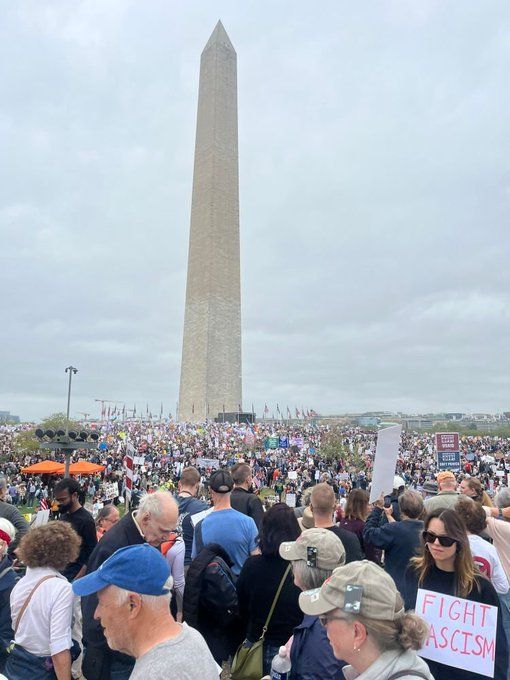 A large crowd gathers near the Washington Monument, holding signs and flags during a protest against fascism under a cloudy sky.
