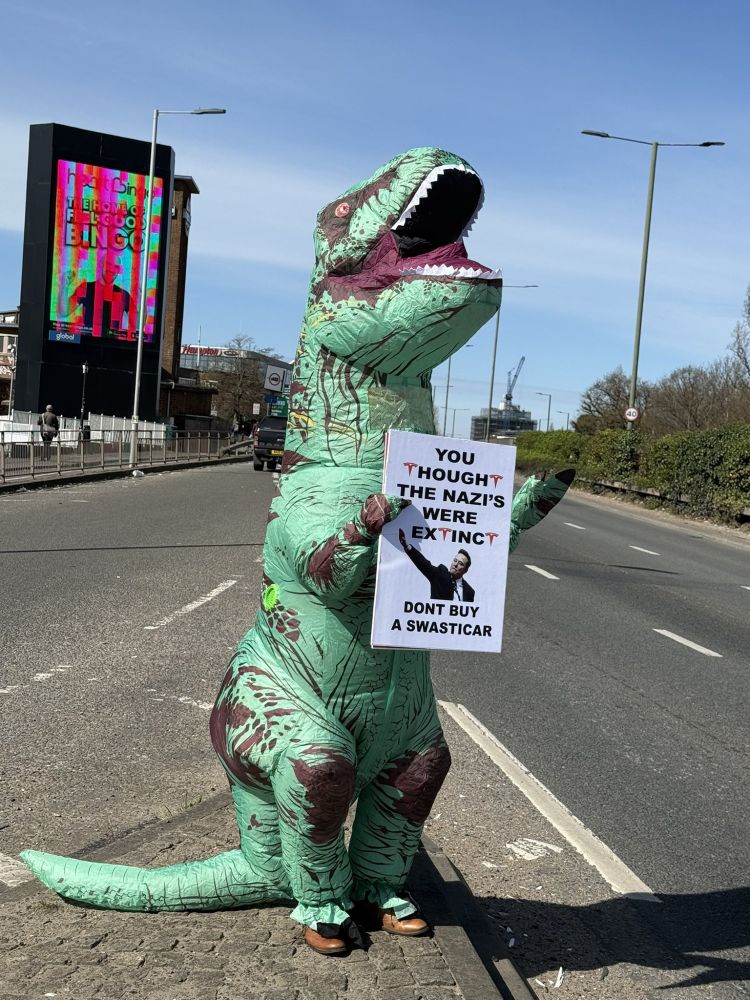 Man in dinosaur suit holds protest sign that says: "YOU THOUGHT THE NAZI'S WERE EXTINCT DONT BUY A SWASTICAR"