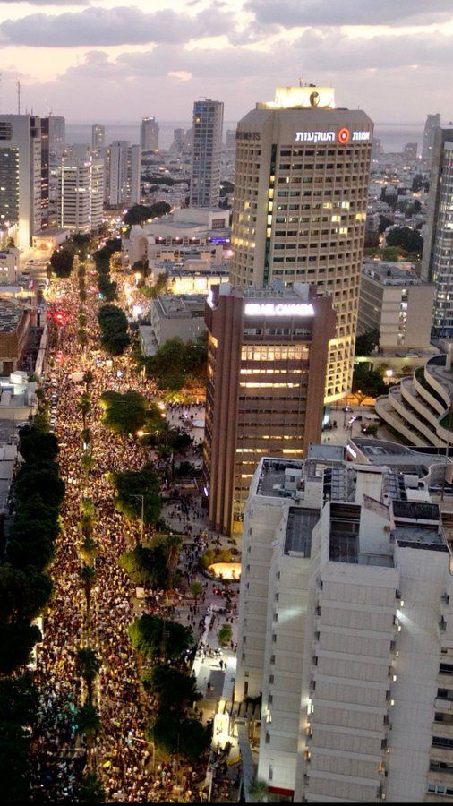 A large Israeli crowd fills dozens of city streets lined with tall buildings during dusk in an urban area.