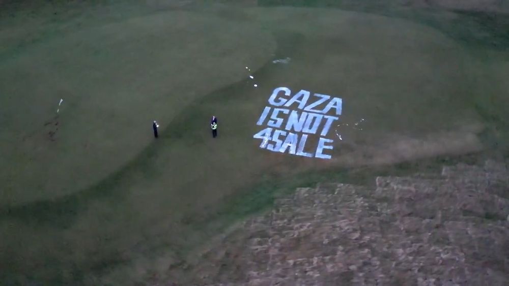 Aerial view of two people standing beside a large painted message on grass reading "GAZA IS NOT 4 SALE."